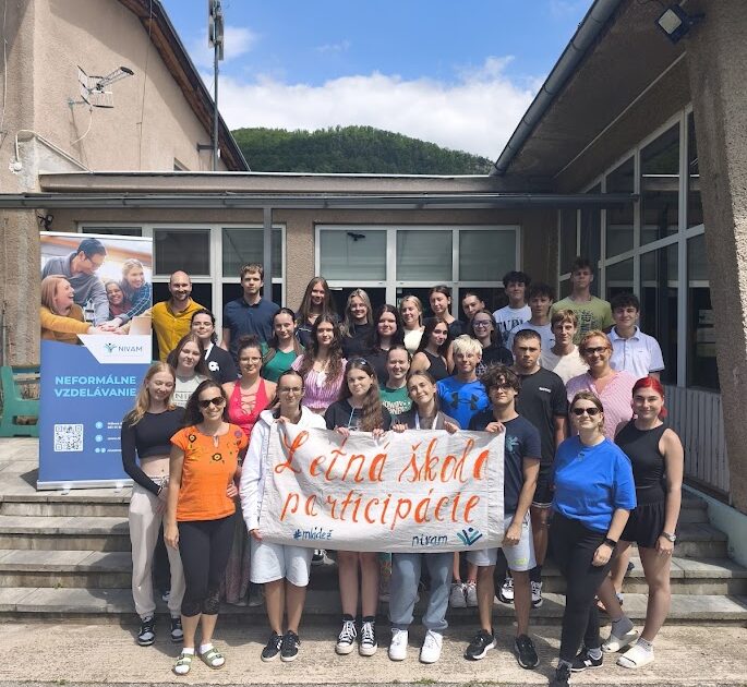Na obrázku stojí skupina mladých ľudí, usmievajú sa do kamery v ruke držia transparent "Letná škola participácie". V pozadí sa nachádza budova a roll up NIVaM inštitúcie